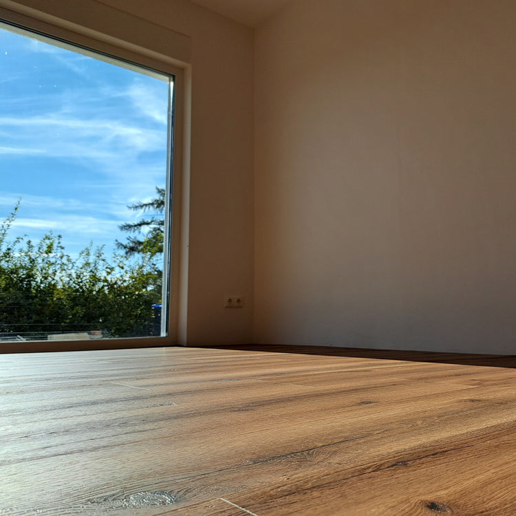 Klick-Vinylboden in Braun in leerem Raum mit weißer Wand und Blick aus deckenhohem Fenster 