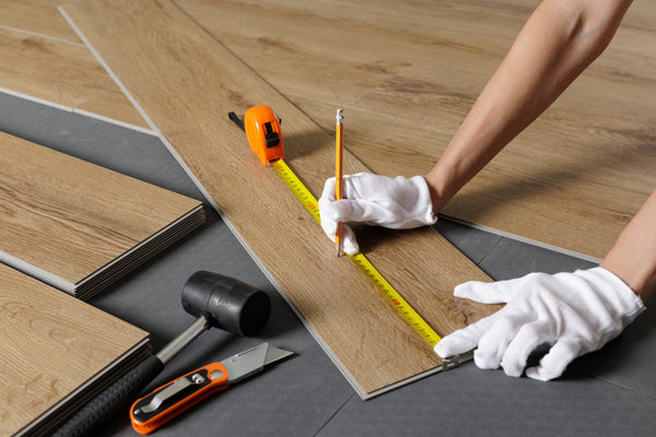 Ein Handwerker der mit einem Maßband ein Stück Klick-Vinyl ausmisst um es zurecht zu schneiden. Es liegt weiteres Werkzeug neben ihm und einige weitere Planken Klick-Vinyl.