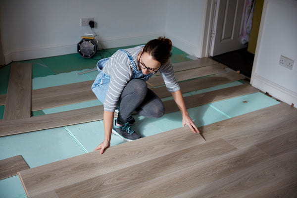 Eine Frau die in ihrem Zuhause Laminat verlegt . Sie kniet auf dem Boden und verlegt eine Planke Laminat. Hinter ihr liegen weiter Planken die noch verlegt werden müssen.